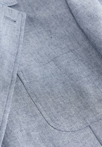 Gros plan sur un tissu de blazer gris clair texturé montrant une poche poitrine cousue et une partie du revers.