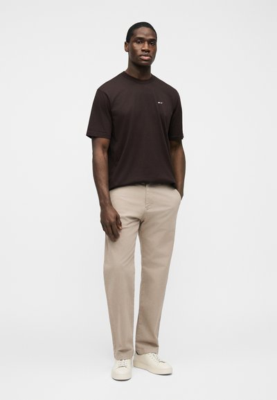 Homme debout, les mains dans les poches, portant un t-shirt marron foncé, un pantalon beige et des baskets blanches sur un fond blanc uni.