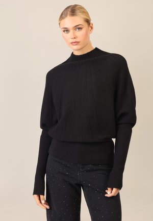 Schwarzer Rippstrickpullover mit hohem Kragen, überschnittenen Schultern und verlängerten Bündchen; getragen mit dunklen Jeans mit hellen Verzierungen.