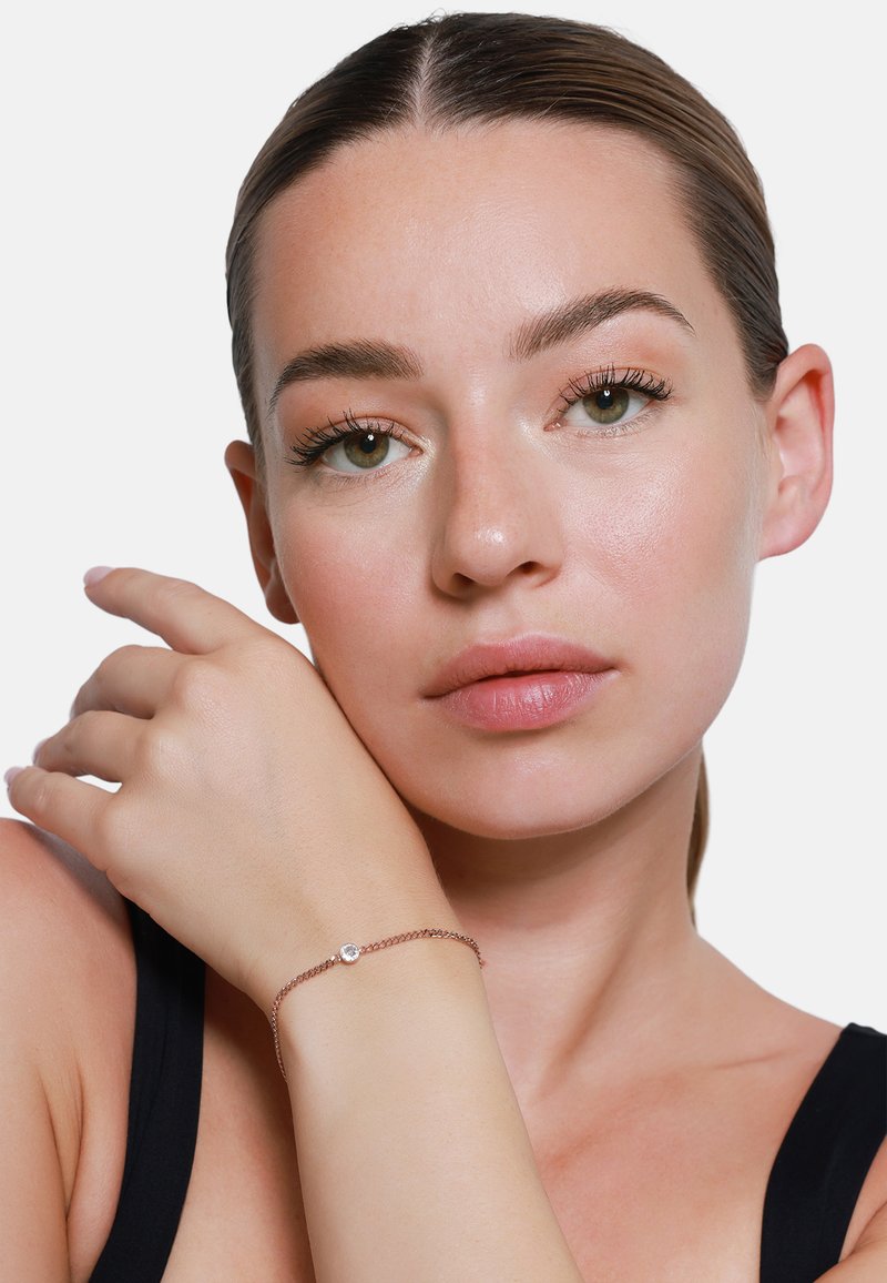 Armband aus Roségold mit zartem Chain, ausgestattet mit einem einzelnen, auffälligen klaren Edelstein, der an einer Hand getragen wird, die am Gesicht ruht.