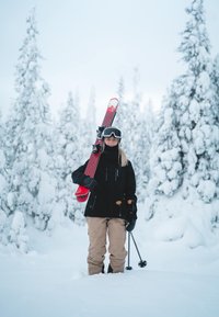 Ski-uitrusting met rood-zwarte ski's vastgehouden door een persoon in een zwart jack, beige broek en handschoenen, staand in een besneeuwd landschap.