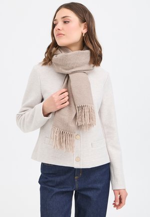 Écharpe en laine beige avec extrémités frangées, portée autour du cou ; associée à une veste gris clair et un jean bleu foncé. Finition texturée.