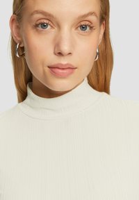 Rippstrick-Turtleneck-Oberteil in Off-White, aus weichem Stoff gefertigt. Das Modell trägt silberne Creolen, hat ein natürliches Make-up und glattes blondes Haar.