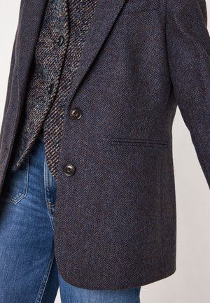 Blazer a motif houndstooth in marrone scuro e blu, caratterizzato da due bottoni, una tasca e una vestibilità tailored, abbinato a jeans blu.