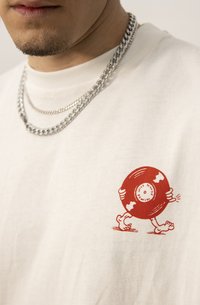 Biely bavlnený tričko s červenou kreslenou grafikou postavy nesúcej vinylovú platňu. Retiazky nosené okolo krku pridávajú detail.