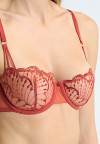 Koronkowy stanik z czerwoną bazą i misternymi haftami kwiatowymi na miseczkach, falbaniaste krawędzie i regulowane ramiączka.