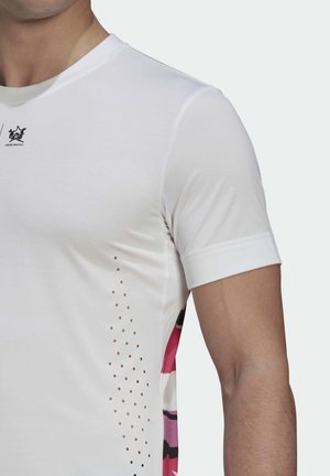 Vit kortärmad skjorta med rund halsringning, med perforerade detaljer på sidan och ett färgstarkt abstrakt mönster längs fållen.