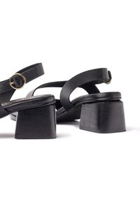 Sandalias de cuero negro con tacón cuadrado, correa de tobillo ajustable con hebilla en tono dorado y diseño de tiras entrelazadas.