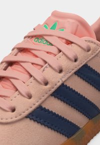 Ανοιχτό ροζ αθλητικό παπούτσι Adidas με ριγέ σε σκοτεινό μπλε, πράσινο λογότυπο στη γλώσσα και ταίριασμα ανοιχτό ροζ κορδόνια σφιχτά δεμένα.