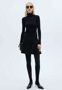 Un col roulé noir à côtes, une jupe mini plissée noire, des collants noirs et des ballerines noires à bout pointu. Des lunettes de soleil en accessoire. Fond simple.