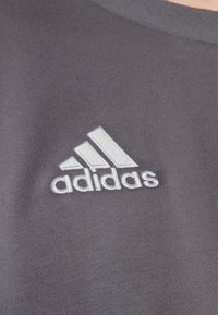 T-shirt de sport gris à manches courtes, col côtelé et logo Adidas brodé en blanc placé sur le côté gauche de la poitrine.