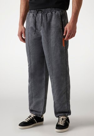 Pantalones de chándal grises con cintura elástica, cordón, bolsillos laterales y un logo de acento naranja, que presentan un corte relajado y una textura suave.