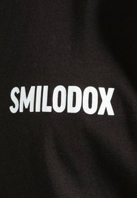 Sort stof med ordet "SMILODOX" i fed hvid skrift, med en struktureret finish, der er typisk for bomulds- eller bomuldsblandingsmaterialer.