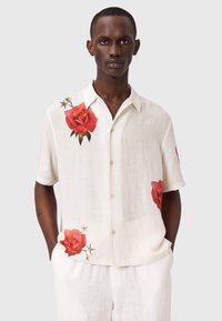 Hombre con camisa blanca de manga corta con estampados de rosas rojas, de pie con las manos en los bolsillos contra un fondo liso.