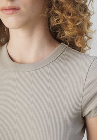 Gros plan d'une personne aux cheveux bouclés châtain clair portant un t-shirt côtelé beige à manches courtes, visible du cou à l'épaule.