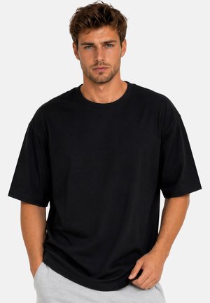 Uomo con capelli castani corti indossa una maglietta nera larga e pantaloni grigio chiaro, in piedi con una mano in tasca su uno sfondo semplice.