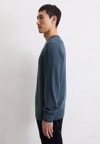 Sweter z dzianiny z wełny w kolorze morskim, z okrągłym dekoltem i ściągaczami przy mankietach. Materiał wydaje się miękki i ma gładką fakturę; noszony razem z czarnymi spodniami.