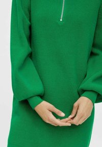 Robe en tricot vert avec un col montant zippé, des manches ballon et une texture côtelée. Les mains sont placées ensemble au niveau de la taille.