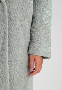 Cappotto grigio chiaro con texture e finitura pelosa. Presenta una tasca laterale e maniche lunghe, evidenziando il materiale morbido e la trama spessa.