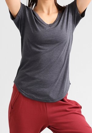 Persoon die een donkergrijze V-hals T-shirt en rode broek draagt, met beide armen boven het hoofd geheven tegen een effen achtergrond.