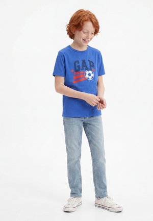 Garçon roux souriant, portant un t-shirt bleu GAP avec un motif de ballon de football, un jean clair et des baskets blanches, debout sur un fond blanc.
