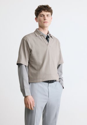 Mladý muž s kučeravými vlasmi, oblečený v sivom tričku s dlhým rukávom pod béžovým krátkym pleteným svetríkom a svetlosivými nohavicami, na bielom pozadí.