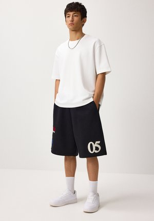 Mladý muž stojí a má na sebe jednoduché biele oversized tričko, čierne šortky s "05" na ľavej nohe, biele ponožky a biele tenisky.