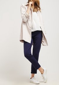 Lichtroze gestructureerde jas met een kraag, witte blouse en navybroek, gecombineerd met witte sneakers. Opvallende knoopdetails op de jas.