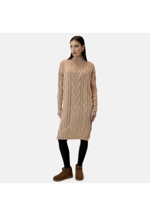 Strikket sweaterkjole i lysebeige med kabelmønster, høj halsudskæring, lange ærmer, ribbet kant og sideslids, kombineret med sorte leggings og brune støvler.