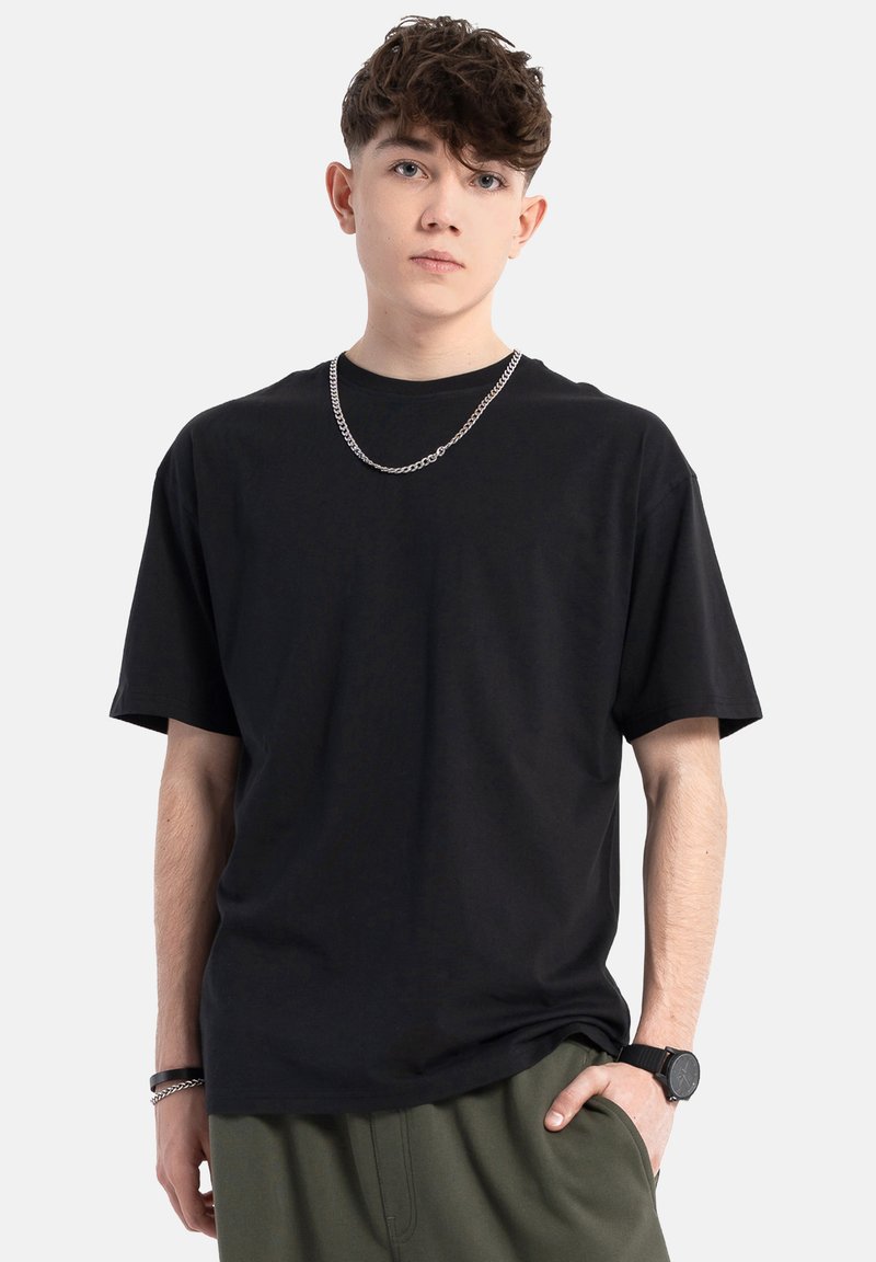 Czarna bawełniana koszulka z krótkim rękawem i okrągłym dekoltem, o luźnym kroju. Noszona z łańcuchem jako dodatkiem oraz oliwkowymi spodniami.