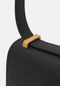 Černý kožený popruh tašky z jemného peblového materiálu připevněný malým obd�élníkovým matným zlatým kovovým prvkem.