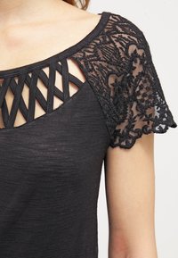 Czarna bluzka z teksturowanej tkaniny z koronkowym detalem na ramionach i wyraźnym dekoltem w kształcie krzyża. Krótkie rękawy z kwiatowymi wzorami.
