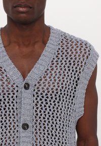 Homme portant un gilet gris en crochet sans manches avec fermeture à boutons sur une poitrine nue, visible du cou jusqu'au milieu du torse.