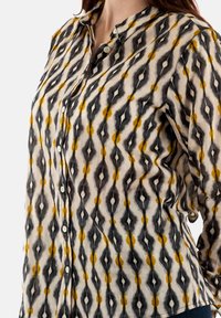 Camisa abotonada hecha de una tela ligera que presenta un patrón ikat en negro, gris y amarillo sobre un fondo crema, con un cuello clásico y botones.