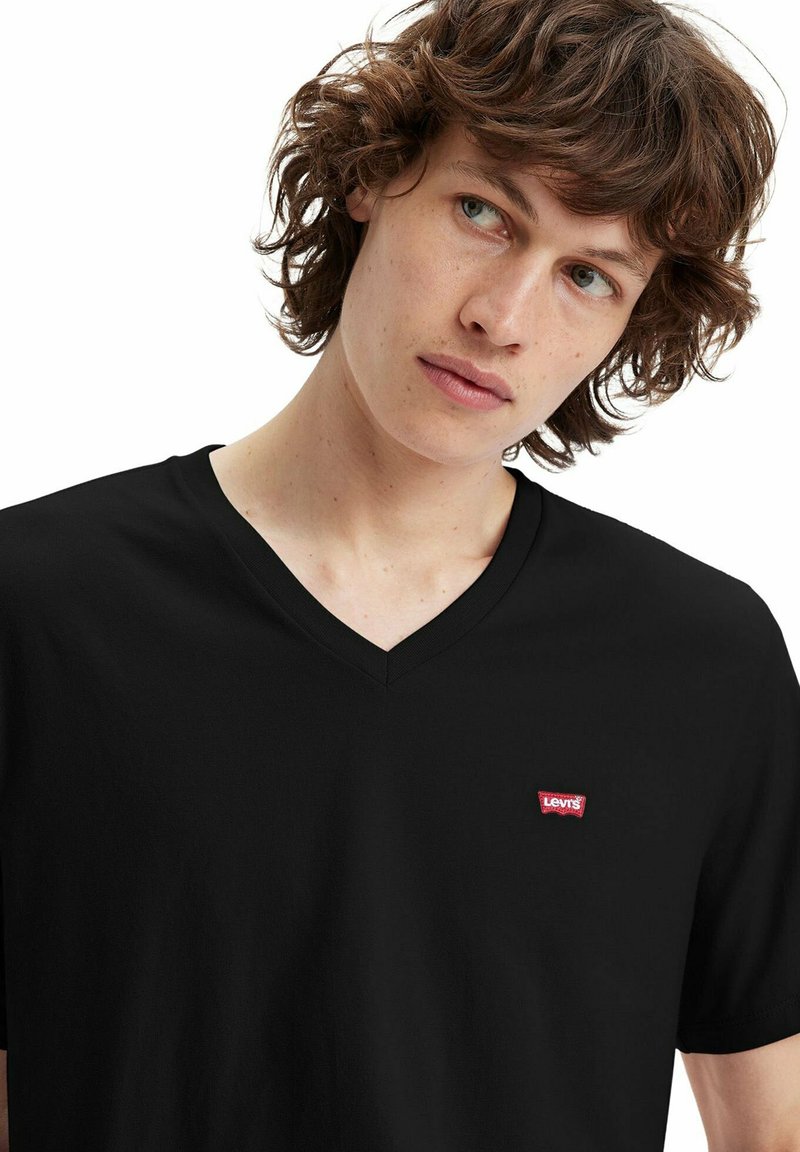 Junger Mann mit lockigem braunem Haar, der ein schwarzes V-Ausschnitt-T-Shirt mit kleinem rotem Levi's-Logo auf der Brust trägt und zur Seite schaut.