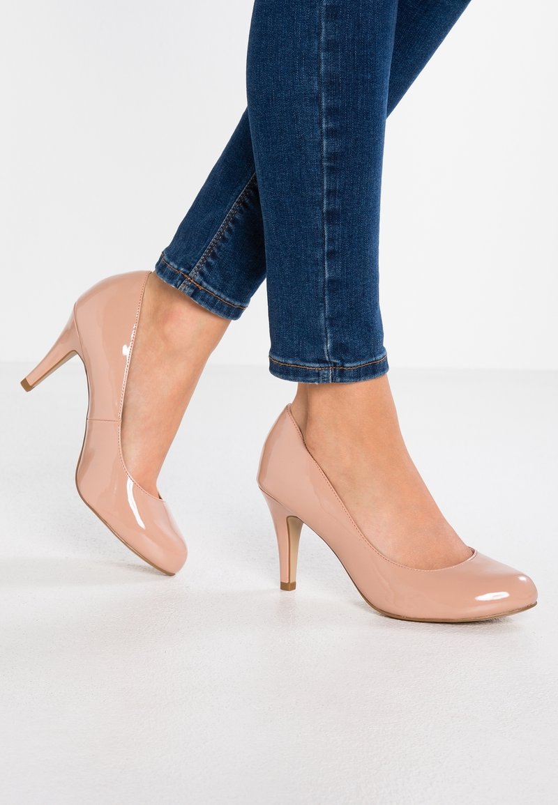 Chaussures à talons hauts en cuir verni couleur nude, avec un bout arrondi, une surface lisse et un talon fin métallique. Portées avec un jean denim ajusté.