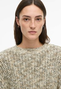 Pull en maille épaisse aux nuances crème, beige et marron ; col rond ; motif texturé. Le mannequin porte des boucles d'oreilles circulaires foncées.