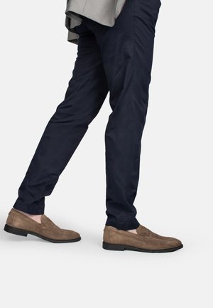 Mann trägt navyblaue Hose und braune Wildleder-Loafer, geht mit einem nach vorne ausgestreckten Bein vor einem einfarbigen Hintergrund.