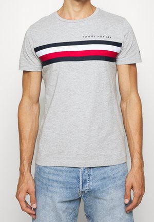Mężczyzna noszący jasnoszary t-shirt Tommy Hilfiger z czerwonymi, białymi i granatowymi poziomymi paskami na wysokości klatki piersiowej oraz jasnoniebieskie dżinsy.