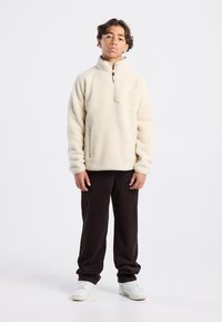 Pullover in pile color crema con zip a un quarto, dotato di un piccolo logo sul petto e due tasche laterali con zip, abbinato a pantaloni in velluto a coste scuri.