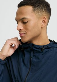 Veste coupe-vent marine avec une finition texturée, dotée d'une fermeture éclair, d'une capuche avec cordon de serrage et de poignets élastiques pour un ajustement réglable.