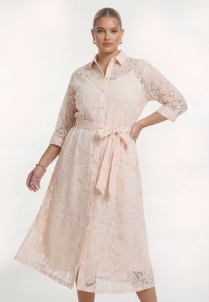 Kvinde iført en lyserød blomsterblonde midi-kjole med 3/4 ærmer, knapper foran og et bælte bundet i taljen, poserer med den ene hånd på hoften.