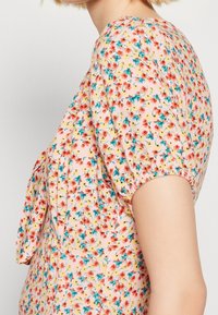 Nainen pukeutuneena haalean vaaleanpunaiseen mekkoon, jossa on pieni punainen ja sininen kukkakuosi sekä puffattuja lyhyitä hihoja, kuvattuna sivusta.