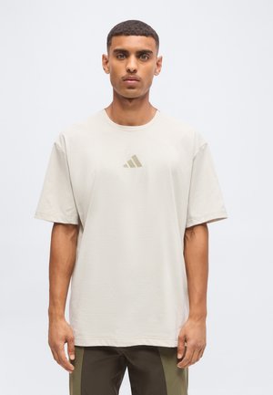 Hombre con el pelo corto que lleva una camiseta amplia de color beige claro con un pequeño logo centrado y pantalones oscuros con paneles laterales.