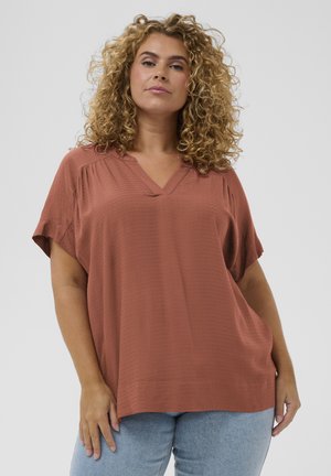 Femme aux cheveux bouclés portant un haut ample à col en V de couleur rouille et un jean bleu clair, debout devant un fond uni clair.
