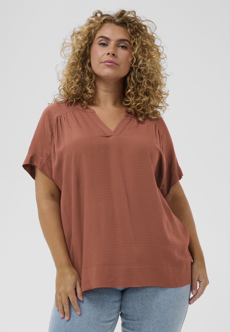 Femme aux cheveux bouclés portant un haut ample à col en V de couleur rouille et un jean bleu clair, debout devant un fond uni clair.