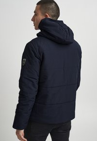 Chaqueta de plumón azul marino con capucha, textura acolchada y un logo bordado en la manga izquierda. Tela durable con un diseño elegante.