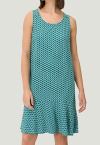 En ærmeløs kjole i teal med et geometrisk mønster af sammenflettede cirkler, med en udvidet kant og glat tekstur.