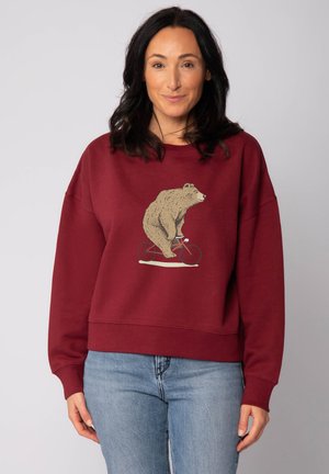 Vrouw in een bordeauxrode sweatshirt met een afbeelding van een beer op een rode fiets, gecombineerd met lichtblauwe jeans, staande tegen een effen achtergrond.