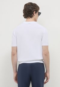Weißes Polohemd mit kurzen Ärmeln und strukturiertem Strickdesign, mit Kragen und gerippten Bündchen, kombiniert mit marineblauen Hosen. Rückansicht gezeigt.